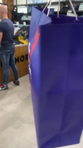 The video opens on a close-up of a large, royal blue shopping bag with the logo of "The Athlete's Foot" printed in white and red. The logo features a stylized red winged foot. As the camera pans slightly, a man in a blue t-shirt and jeans is visible in the background, standing at a counter in a retail store. The store appears to be a clothing or shoe store, with shelves stocked with merchandise in the background. The lighting is bright and the environment seems to be indoors. The video captures the shopping bag from different angles, with a slight blur indicating movement.