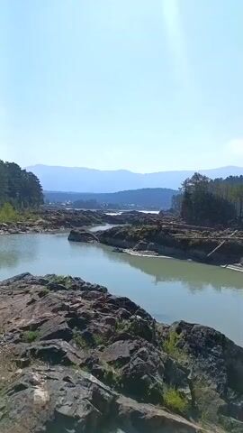 A scenic river flows through a rocky landscape. The water is a light green color, and there are several large rocks and boulders in the foreground and midground. The riverbanks are lined with trees, and in the distance, there are mountains. The sky is clear and blue, with the sun shining brightly. The video appears to have been filmed on a clear, sunny day. There are no people or animals visible in the video.