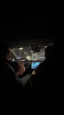 The video shows the interior of a car at night. Two people are visible from behind, seated in the front seats. The driver, on the left, is holding the steering wheel and appears to be wearing a pink shirt and a hat. A large screen is mounted in the center of the dashboard displaying a navigation map. The passenger on the right is also looking towards the screen. Outside the car, lights from streetlights and other vehicles are visible, indicating a road environment, possibly a toll booth or checkpoint, with multiple cars in front. The scene is dimly lit, with the primary light sources being the car's dashboard screen and the exterior lights.