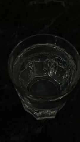 A close-up, high-angle shot shows a slice of lemon being dropped into a glass of water. The lemon slice, which is round with a yellow rind and greenish-yellow pulp, is seen descending into the clear liquid. The glass is made of clear glass and has a faceted base. The background is dark and appears to be a marbled surface. The video is short, lasting only a few seconds.