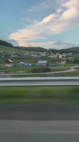 The video shows a blurry view from a moving vehicle of a rural village nestled in a valley with rolling hills covered in trees and grass. A white metal guardrail runs horizontally across the lower portion of the frame, and the foreground is blurred green grass, suggesting motion. In the middle ground, there are numerous small houses with colorful roofs, interspersed with patches of green fields and trees. A few tall, thin utility poles with wires are visible. The sky is blue with scattered white clouds, some tinged with pink, indicating it might be late afternoon or early evening. The overall impression is a peaceful, scenic rural landscape observed from a moving vehicle.