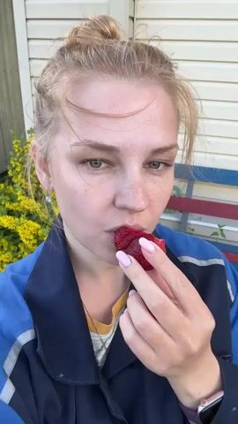The video shows a young woman with blonde hair pulled back in a bun, eating a strawberry. She is wearing a blue work jacket over a striped shirt. The woman has freckles and is looking down at the strawberry as she bites into it. Her fingernails are painted a light pink.

The background suggests an outdoor setting, with a beige siding house and some yellow flowers visible. A bench with red and blue stripes is also present. The lighting indicates it is daytime, likely sunny. The woman appears relaxed and is enjoying her snack. There is no visible text in the video. The location context provided is Дунилово, Russia.
