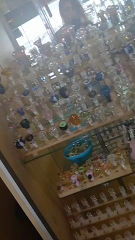 The video shows a display case filled with glass bowls, presumably for smoking. The bowls are arranged on multiple shelves, with various colors and designs. A person is reflected in the glass of the display case, but they are not clearly visible. The environment appears to be a retail setting, possibly a smoke shop or a head shop. There are no discernible actions or interactions taking place in the video.