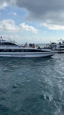 A white passenger boat named "AXE STONE" and "AXE STONE WARRIOR" is the central focus, appearing to be either stationary or moving slowly on choppy blue-green water. The boat also has "GOLDEN MOUNTAIN" written on its side, along with contact information: "+62 8123 7623 130" and "axestonetravel@gmail.com". Several people are visible on the upper deck of the boat. In the background, other boats are partially visible, and a distant landmass can be seen under a partly cloudy sky. The scene is set outdoors during the daytime in Kampung Toyapakeh, Indonesia.