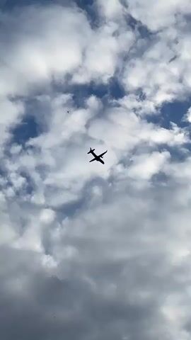 The video shows an airplane flying in a cloudy sky. The plane is small in the frame and appears silhouetted against the clouds and blue sky. The clouds are a mix of white and gray, suggesting varied lighting conditions. The overall view is upwards, with the camera pointed towards the sky.