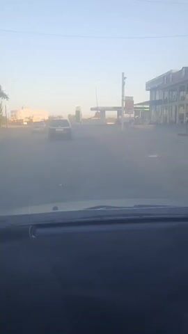 The video shows a view from inside a vehicle, looking forward. The time stamp indicates the beginning of the recording at 00:00. The scene appears to be an outdoor environment, possibly a road or street. In the distance, there is a gas station with visible pumps and a canopy. There are also buildings on the left and right sides of the road. Several cars are present on the road, some moving and some stationary. The sky is visible and appears clear, suggesting it is daytime. The image quality is somewhat hazy, possibly due to dust or a dirty windshield. The video continues to play, showing the vehicle moving forward and the gas station and surrounding environment remaining in view. No people are clearly visible in the video. The focus is on the road ahead and the elements within the visible landscape.