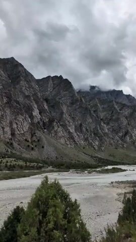 This media file captures a vast, rugged mountain landscape under a cloudy sky. The mountains are steep and rocky, with visible strata and some patches of snow near their peaks. The slopes are sparsely populated with trees, creating a stark contrast between the barren rock and the sparse vegetation.

In the foreground, a wide, shallow riverbed dominates the scene. The river itself appears to be a calm, grayish-blue, with a wide expanse of gravel and sand banks on either side. A few evergreen trees are scattered along the riverbanks, adding touches of green to the otherwise muted color palette. The overall impression is one of a remote, natural, and somewhat desolate environment.

The time of day appears to be overcast, likely mid-day or late afternoon, given the diffused light and the absence of strong shadows. The weather is cloudy, contributing to a somber and dramatic atmosphere. The location cues, such as the type of vegetation and the geological formations, along with the provided context of Darcha, India, suggest a high-altitude, mountainous region.

There are no people or specific objects prominently featured in the scene, with the focus being entirely on the natural landscape. The activity, if any, is the gentle flow of the river through the valley. The emotions evoked are likely awe, solitude, and perhaps a sense of the immense power and scale of nature. No visible text is present in the media file.