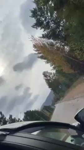 The video shows a first-person perspective from inside a car driving on a road. The car is moving alongside trees and a mountain can be seen in the distance. The sky is overcast with clouds.