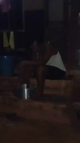 A person is seated on concrete steps in the dark. They are wearing a white tank top and dark shorts, and are barefoot. A metal pot is placed on the step in front of them, and their feet are inside it. Their head is bowed, and they rest an arm on their knee. A blue bucket is visible to the left of the frame, and laundry is hanging on a clothesline in the background. The lighting is poor, with the scene appearing to be poorly lit.
