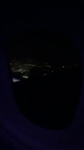 The video shows an aerial view of a city at night, as seen through the window of an airplane. The city is illuminated by numerous lights, forming a network of roads and buildings. The aircraft wing is partially visible in the lower left corner of the window. The surrounding sky is dark.