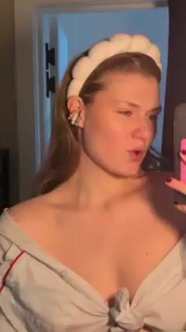 A woman with fair skin and long hair is in a dimly lit room. She wears a white, button-down shirt tied at the waist and a white headband. She is holding a cellphone with a pink, grid-patterned grip in her right hand, using it to record herself in a mirror. Her left hand is initially outstretched towards the camera.  She then touches her neck and face. The room has a white door with dark hardware and dark curtains. Music plays in the background.