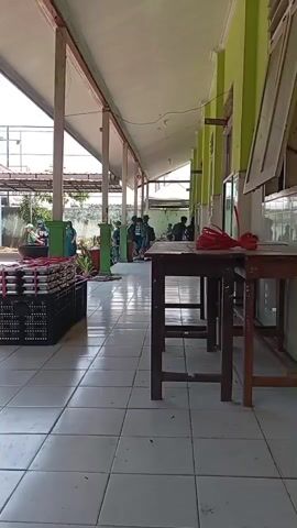 A long, covered walkway with green walls and white tiled floors is shown. Several people, mostly young men, are standing and walking in the walkway. There are stacks of black crates filled with items on the left side of the walkway. There are also two wooden tables in the foreground on the right side of the walkway. The sky is visible in the background, and it appears to be a sunny day. The overall impression is that this is a school or a similar institution.
