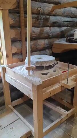 The video displays the interior of a mill, likely a historical one. The structure is made primarily of wood, with exposed beams and log walls. In the foreground, a wooden platform and railing are visible. Suspended from an overhead beam is a heavy, black metal tool, possibly a lifting or gripping mechanism, with two curved arms that end in hooks. Behind this, a large wooden chest or bin sits on the floor. The background reveals more milling equipment, including sacks of grain, various wooden structures, and bundles of wheat stalks or other grains used for decoration or display. The lighting appears to be natural and ambient. There are no people visible in the video. The overall impression is of a preserved historical industrial space.