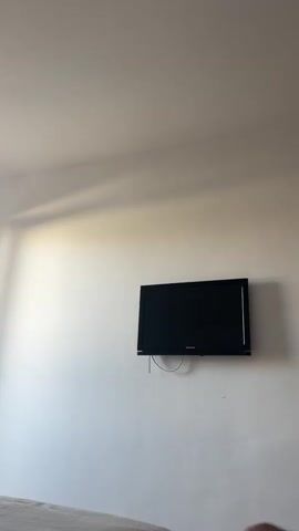 A TV is mounted on a white wall. A window with a white frame is partially open, with curtains on the left side. Sunlight streams in from the window, casting shadows on the wall. A bed is visible in the foreground. The room appears to be a bedroom.