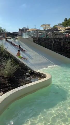 The video shows a group of children and young people at a water park. They are sliding down a water slide into a shallow pool. The water slide is white and appears to be made of plastic. The pool is filled with clear, turquoise water. There are lounge chairs and umbrellas in the background, suggesting a resort or hotel setting. The sun is shining and the sky is clear blue. The people are wearing swimwear and appear to be enjoying themselves. One person is on a blue inflatable ring, and others are sliding directly on the slide. The action progresses as more people slide down, with the first person reaching the pool and splashing.