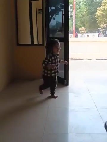 A young child in a black and white checkered shirt and dark pants walks across a tiled floor. The child has dark hair and is barefoot. The child is moving towards a doorway. Another child, wearing an orange long-sleeved shirt and patterned pants, enters the frame from the right, walking in the same direction as the first child. A green plastic chair is visible in the background, to the left of the doorway. A dark wooden table or furniture piece is partially visible in the foreground, bottom right. The scene appears to be indoors, possibly a hallway or entrance area.