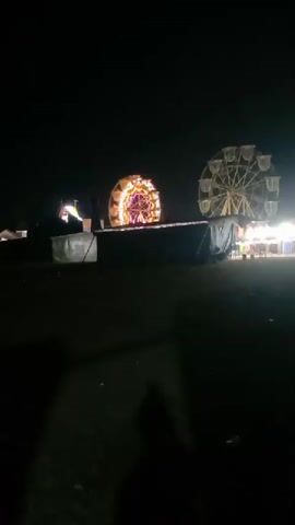 The video shows a nighttime scene at what appears to be a carnival or fair. Two Ferris wheels are visible, one larger and closer to the viewer, and another smaller one further in the background. There are also several tents or stalls set up, illuminated by lights. In the background, a tall tower-like structure, possibly a cell phone tower, is silhouetted against the dark sky. The ground in the foreground is dark and appears to be uneven, with some scattered debris. The overall atmosphere is that of a lively event taking place after dark, with the rides and stalls brightly lit.