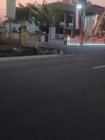 A video captured at dusk shows a street in front of a building decorated with Indonesian flags and red and white lights. Several motorcycles are seen driving on the asphalt road. The building appears to be a commercial establishment or a public facility. The overall atmosphere suggests a festive or commemorative event.