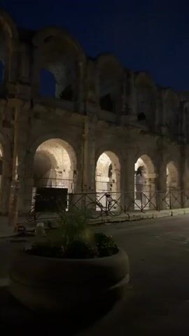 The video shows a nighttime view of a large, ancient stone structure with arches. The structure is illuminated, with warm light shining through the arches and casting shadows. In the foreground, there is a dark area with a planter containing some greenery. A street runs in front of the structure, and a fence separates it from the building. A bicycle is visible behind the fence. Further in the background, some people are walking, and there are lights from other buildings and possibly streetlights. The sky is dark blue, indicating it is evening or night. The video is somewhat shaky and the focus shifts, making some details blurry, especially towards the end.