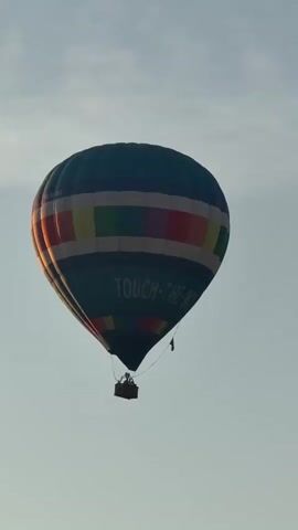 A hot air balloon with blue, yellow, red, and green squares is seen flying in a pale blue sky with some clouds. The balloon has text that reads "TOUCH-THE-SKY". Below the balloon is a basket with two people visible. The camera is positioned below and looking up at the balloon.