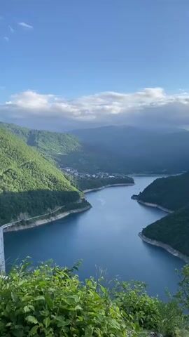 The video captures a panoramic view of a serene blue lake (Pivsko Jezero) winding through majestic, verdant mountains under a bright blue sky dotted with scattered white clouds. Lush green foliage frames the foreground of the shot. A long, white bridge stretches across the lake on the left side of the frame, and a small town or village is visible nestled in the valley on the far side. The sunlight illuminates the landscape, casting shadows on the mountain slopes, suggesting it is daytime. The scene is tranquil and devoid of people, showcasing the natural beauty of the area near Plužine, Montenegro. No text is visible.