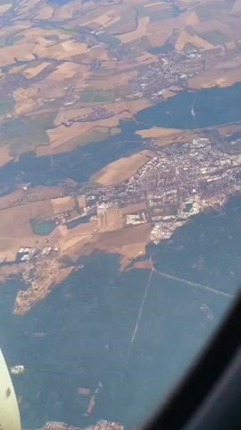 A high-angle aerial view shows a town nestled between agricultural fields and dense forests. The town appears to be a cluster of buildings with a network of roads running through it. The surrounding landscape is a patchwork of tan-colored fields and dark green forested areas. A body of water, possibly a lake or pond, is visible to the left of the town. Part of an airplane wing is in the lower left corner of the frame. The sun is casting shadows, indicating it is daytime.