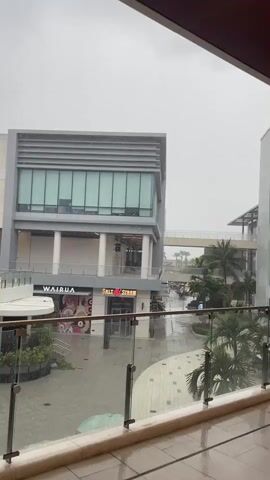 The video pans across a covered outdoor area of what appears to be a shopping mall or modern building complex on a rainy day. In the foreground, a glass railing with metal posts is visible, suggesting a balcony or walkway. The ground outside is wet and reflects the overcast sky.

In the midground, several buildings with modern architectural designs are visible. One building features a light-colored stone facade on the upper section and a gray section with horizontal window elements on the lower portion. Below this, the storefronts of two businesses are apparent: "WAIRUA" with an image of donuts and another with a red sign reading "SALT & STRAW". There are covered walkways and pillars supporting the upper levels of the structures. Tropical trees, such as palm trees, are interspersed throughout the outdoor area.

In the background, more buildings and some distant trees can be seen under the gray, rainy sky. The rain is visible as streaks falling through the air, particularly noticeable in the ambient light. There are no people clearly visible in the frame, and no specific actions or interactions are taking place. The overall atmosphere is calm and slightly somber due to the weather.