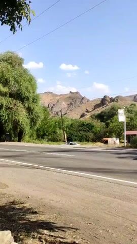 A black car drives down a two-lane road. The road is flanked by dry, dusty terrain and green trees. In the background, there are rocky hills and a clear blue sky with some white clouds. A white pole stands near the road.