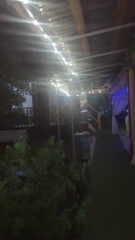 This media file captures an outdoor evening scene under a covered patio adorned with string lights. The setting appears to be a backyard or a similar domestic outdoor space, possibly in Alakhadzi, Abkhazia. The time of day is clearly night, indicated by the darkness and the artificial illumination from the string lights and interior lights of a building visible in the background.

The main subjects are the setting itself and the atmospheric lighting. There are no visible people in the frame. Objects present include a corrugated metal roof supported by wooden beams and metal poles, a railing, some greenery in the foreground, and what looks like patio furniture. In the background, a curtained doorway and a wooden staircase suggest the entrance to a building or another part of the property. The lighting creates a festive and somewhat magical ambiance, with the bright bulbs casting long streaks of light and reflections on the wet or shiny surfaces of the roof. The overall mood is peaceful and inviting, evoking a sense of quiet enjoyment or anticipation. There is no visible text in the media file.