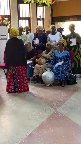 A group of women in traditional African attire are gathered indoors, seemingly in a community hall or church.  They are singing and some are holding papers, likely lyrics. In the foreground, a woman in a red and black dress faces away from the camera, while other women are seated and standing behind her.  One woman on the left is playing a drum.  There is a large silver pot in the center of the seated women. The room has a tiled floor and windows in the background.