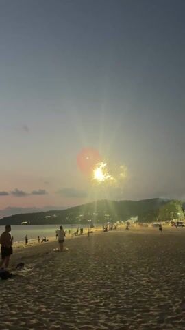 The video captures a beach scene at dusk with a series of fireworks going off in the sky. The sky is a blend of dark blue and light pink hues. In the background, a land mass with trees and scattered lights along a ridge is visible.  The beach is sandy, and several people are strolling along the shore, standing, or sitting.  A few structures that appear to be stalls or tents are set up on the far right side of the beach, illuminated by artificial lights. The fireworks are bursting above the scene, initially as clusters of red lights, then as golden starbursts.