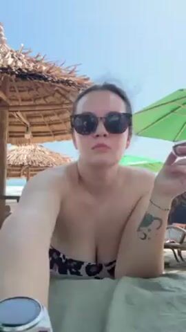 A woman with sunglasses and a cheetah-print strapless top is eating something on the beach. She has a watch on her left wrist and a tattoo on her right. The background shows a sandy beach with beach umbrellas, some thatched and some green. The sky is clear and blue.