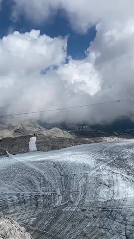 The video shows a high-angle view of a glacier with visible striations, indicating the movement of ice. A cable car line stretches across the frame, with two small red gondolas visible in the distance. Rocky mountains rise on the left and in the background, with a prominent peak on the right. The sky is partly cloudy with patches of blue. The scene appears to be in a mountainous, alpine environment.