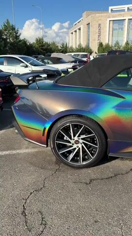 A convertible car with a holographic wrap is parked on asphalt. The car is grey with a rainbow effect that shifts colors. The top is down, revealing a black interior. The car has black wheels with red accents. In the background, other cars are parked, including a lime green convertible. A building with windows and a sign reading "NOVICOTECH" is visible behind the cars. Trees and a blue sky with clouds are also present. The video pans across the car from the rear to the front.