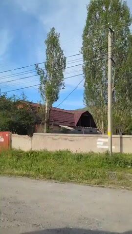The camera moves along a dirt road, passing a house with a red roof behind a concrete fence. There are utility poles and wires above. The sky is blue with clouds. A sign on a red metal structure is visible briefly, with text in a different language. Grass and trees line the road. The camera appears to be inside a moving vehicle, indicated by the occasional view of the side mirror.