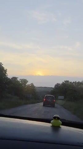 A dashcam view of a car driving on a road toward a sunset, with a toy duck with sunglasses on the dashboard. Another car is ahead on the road.