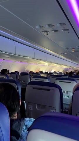 The video shows the interior of an airplane cabin, viewed from a seat towards the back. The seats are predominantly blue and gray, and most are occupied by passengers. Overhead, purple LED strip lights run along the ceiling. The cabin is well-lit with overhead lights. The video appears to be taken before takeoff or during a flight. There is a focus on the seats and the general atmosphere inside the aircraft.