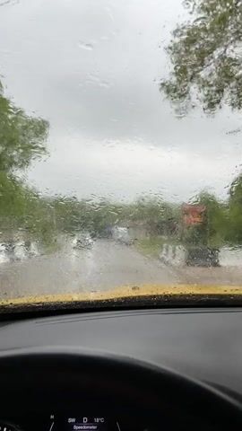 The video shows the view from inside a yellow car driving on a rainy day. The windshield wipers are on, clearing the raindrops. Outside, the street is wet, with parked cars and trees visible. The car's dashboard shows a temperature of 17-18 degrees Celsius.