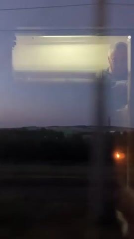 The video shows a man with glasses sitting inside a train compartment. He is looking down and appears to be holding something in his hands, possibly a phone or a book. The train is moving at dusk, and the outside scenery is dark with some lights visible in the distance. The compartment is illuminated by an overhead light.