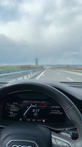 The camera is mounted behind the steering wheel of a car, looking out through the windshield at a multilane highway. The car is moving at high speed, indicated by the dashboard display fluctuating between 217 and 223 kilometers per hour. The dashboard also shows the time as 12:40, an outside temperature of 13.5 degrees Celsius, and fuel consumption of 11.6 liters per 100 kilometers. A head-up display projects speed onto the windshield.  Ahead, the highway stretches into the distance under a cloudy sky, with several other vehicles including a large white truck in the right lane. Guardrails line both sides of the road. The car's dashboard also displays a call list with names such as "Dad," "Mimet," "Bosa," and "Mum."