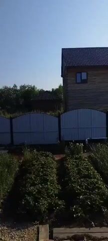 The video shows a rural backyard on a sunny day. The camera pans across a garden with rows of green plants, a white bathtub filled with water, and a large greenhouse. Several buckets and containers are also visible near the greenhouse. In the background, there is a wooden house and a fence. The overall impression is of a functional, lived-in space.