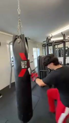 A young man is shown in a gym, wearing a black t-shirt, red pants with white stripes, and red and black boxing gloves. He is punching a tall, black punching bag with the words "MOVE BETTER" printed on it in red and white. The punching bag is suspended from the ceiling by a silver chain. The gym has black flooring and various weightlifting machines in the background. There is also a window visible in the background.