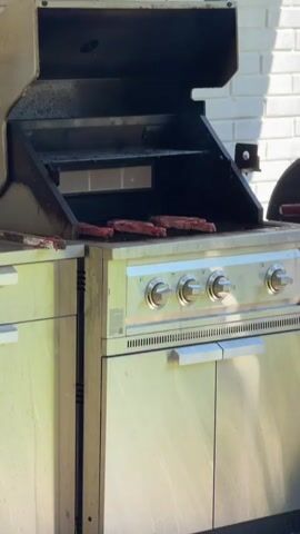A static medium shot captures a stainless steel outdoor gas grill with its lid open. Four pieces of raw, red meat are visible on the grill grates, positioned towards the center-right of the cooking surface. A pair of metal tongs rests on the left side shelf of the grill, partially obscured. The grill features four control knobs on its front panel and a lower cabinet with two doors. A white brick wall is visible in the background, illuminated by sunlight.