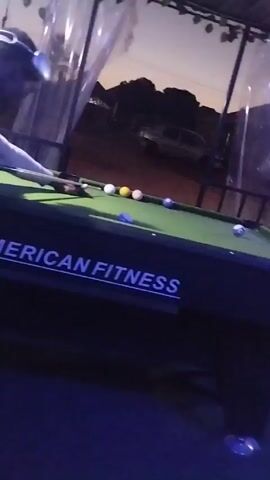 The video shows a person playing pool on a table labeled "AMERICAN FITNESS". The lighting changes throughout the video, shifting from green to blue to red, then returning to green. The pool table has several balls on it. The person playing is wearing a light-colored shirt. The pool table appears to be outdoors under a canopy. A car is visible in the background. Music plays throughout the video.