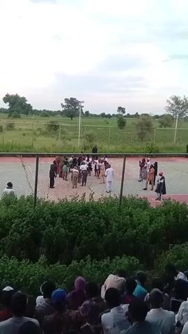 A group of people are gathered on an outdoor court with a net. The court appears to be a tennis or basketball court. The people are dressed in a variety of clothing, some in traditional attire and others in casual wear. Some of the individuals are engaged in conversation or observing an activity on the court. In the background, there is a grassy field with trees and a cloudy sky. A fence surrounds the court. In the foreground, there is a crowd of people sitting and watching the scene on the court. The overall impression is of a community gathering or event taking place outdoors.