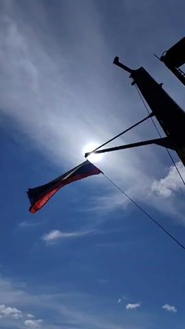 The video shows a Russian flag fluttering against a clear blue sky partially obscured by thin white clouds. The sun shines brightly behind the flag, and the silhouette of a mast and rigging are visible. The flag, appearing weathered, is attached to the mast with a thin line. The perspective is a low-angle shot, emphasizing the flag against the sky. There is the sound of an engine.