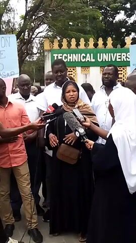 In a medium shot, a woman wearing a black abaya and a tan headscarf speaks into several microphones, surrounded by a group of people, many of whom appear to be listening attentively. She's also carrying a small brown purse on her shoulder. The background features a green sign that reads "TECHNICAL UNIVERSITY" in white capital letters. Some individuals hold signs; one visible sign reads "MILLION BE USED ASAP". The people present include men in casual attire such as white shirts and a salmon-colored shirt, and another woman wearing a white hijab. The setting appears to be outdoors, near what looks like the entrance to the university. The atmosphere suggests a press conference or a public statement being made amidst some sort of gathering.