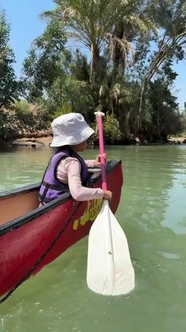 A child wearing a white bucket hat and a purple life vest sits in a red canoe on green water, holding a paddle with a pink shaft and a white blade. The child is facing away from the camera, and the paddle is in the water. Trees and vegetation are visible on the bank in the background, with palm trees being prominent. The sky is blue and clear. There appears to be some text on the side of the canoe. The video shows the child paddling.