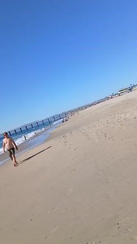 The video shows a sunny day at a beach. The sky is clear and blue. The sand is light brown. A pier stretches out into the ocean, with people walking on it. Several people are wading in the water near the pier. A man in shorts and sunglasses is walking along the beach in the foreground. The waves are small and white. The beach appears moderately populated.