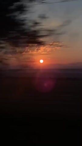 The video shows a sunset with a clear sky and some clouds. The sun is visible as a bright orange orb. The scene appears to be a landscape with fields and trees. The video is shaky and appears to have been filmed from a moving vehicle.